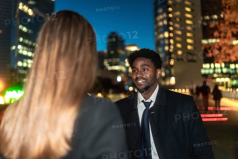 Young african american man engaging in a business conversation with a female colleague outdoors, with blurred city lights and buildings providing a dynamic urban backdrop during the evening
