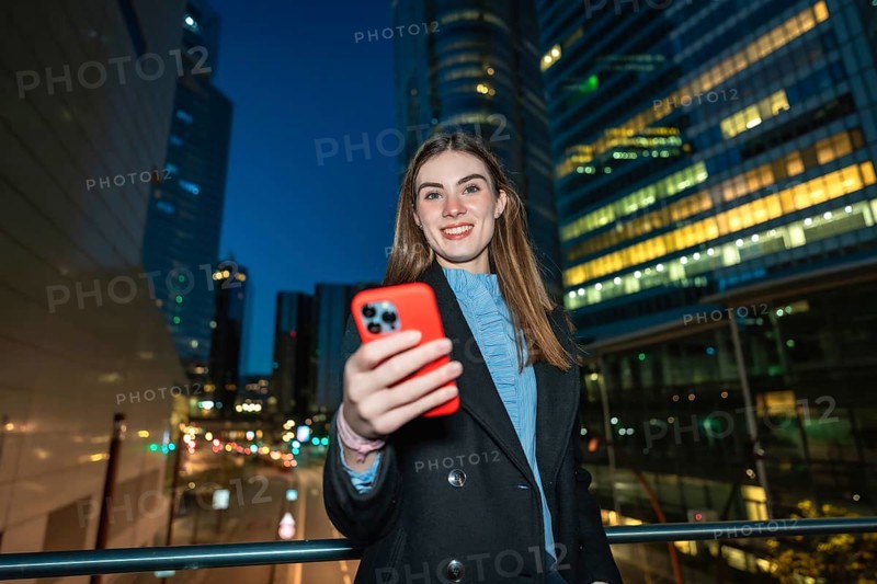 Young woman smiling and holding a smartphone, capturing a selfie or video while standing on a bridge in a modern city at night with illuminated skyscrapers in the background