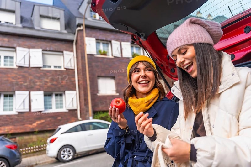 Two cheerful young multiracial women enjoying a moment of laughter while unloading fresh produce, including a red apple, from a car trunk after a successful grocery shopping trip