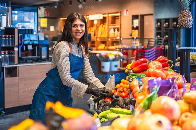 Woman in apron and gloves arranging fresh avocados on a supermarket produce display, smiling while organizing fruits and vegetables in a modern grocery store setting