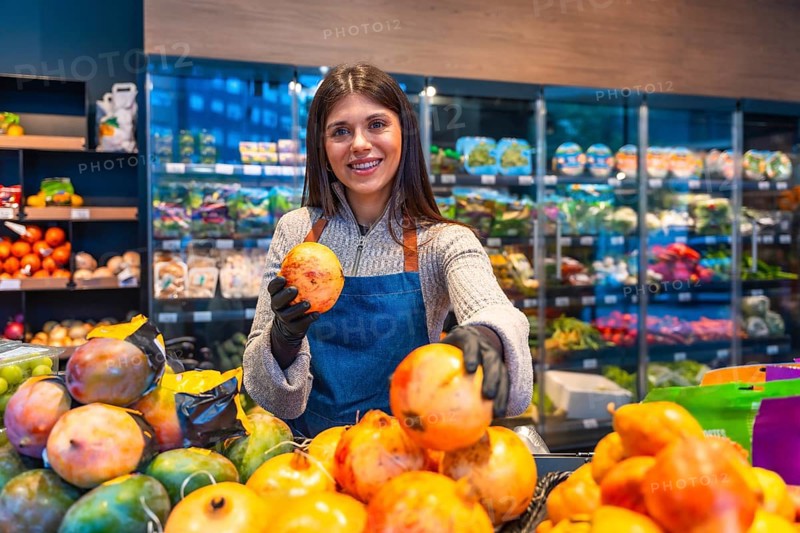 Smiling young woman greengrocer wearing an apron and gloves at a supermarket produce stand, proudly presenting fresh pomegranates to customers, ensuring quality and excellent service