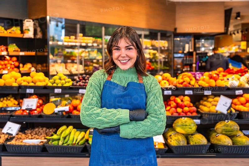 Smiling young greengrocer in blue apron and green sweater stands confidently with arms crossed amid abundant colorful fruits and vegetables in a supermarket produce aisle