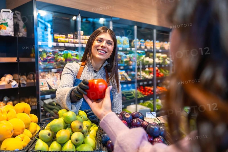 Friendly saleswoman in apron and gloves offering a fresh red bell pepper to a smiling customer in a bright supermarket produce section, highlighting helpful service and healthy shopping