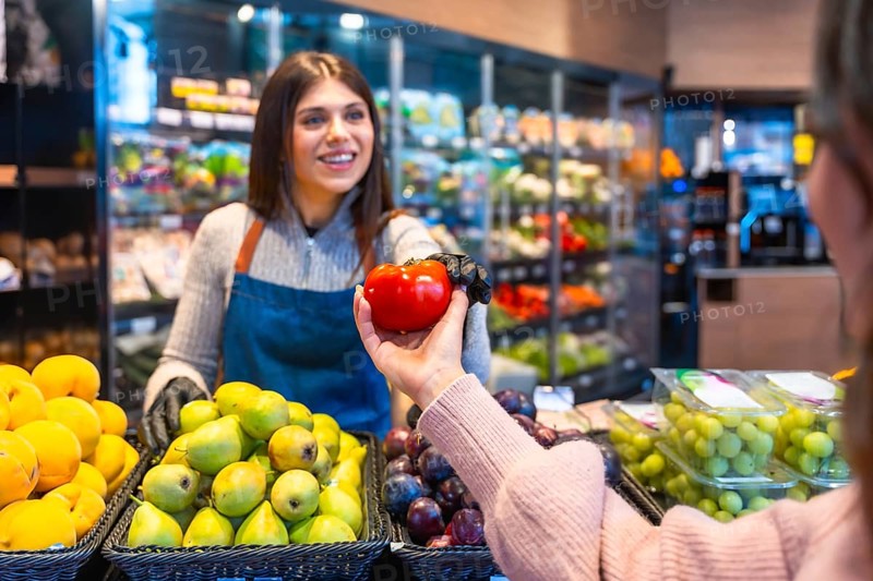 Greengrocer handing a ripe red tomato to a customer during grocery shopping in a produce section, showing excellent store service and healthy food choices