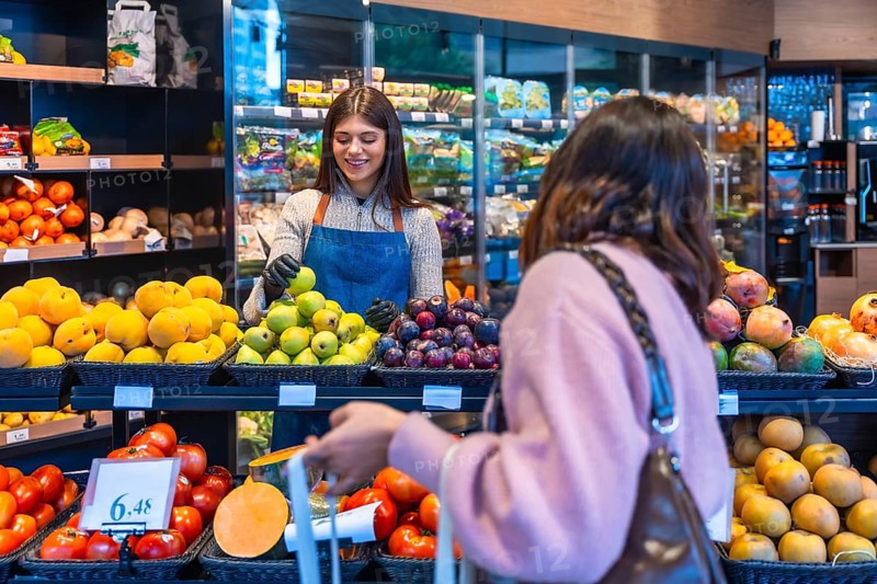 Friendly supermarket employee wearing an apron and gloves assisting a female customer in the produce section by offering a fresh green apple, highlighting good service and healthy food choices