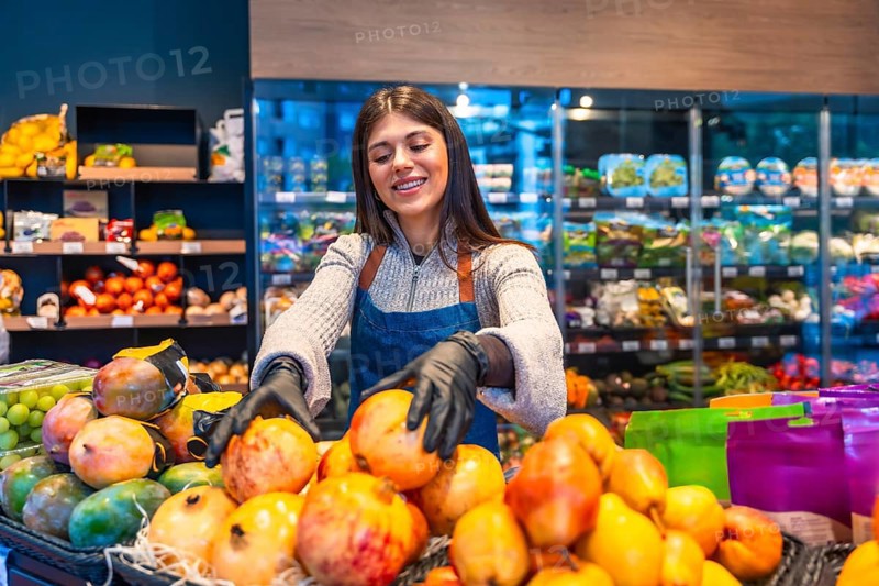 Smiling young supermarket employee in apron and gloves arranging fresh pomegranates, mangos and grapes on produce display, highlighting quality, care and healthy retail service