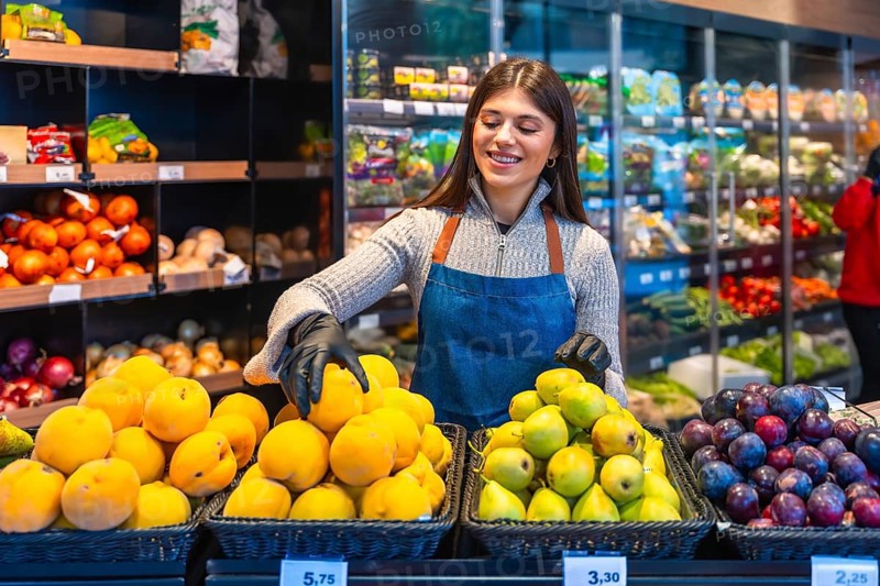 Woman grocer wearing gloves and an apron, smiling while arranging fresh produce like apricots, pears, and plums on display in a modern supermarket or grocery store