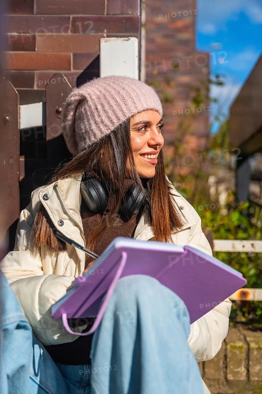 Young woman in knitted hat and headphones smiling while drawing on a digital tablet outdoors in autumn sunlight, cozy and focused, enjoying creative work in a park like urban setting