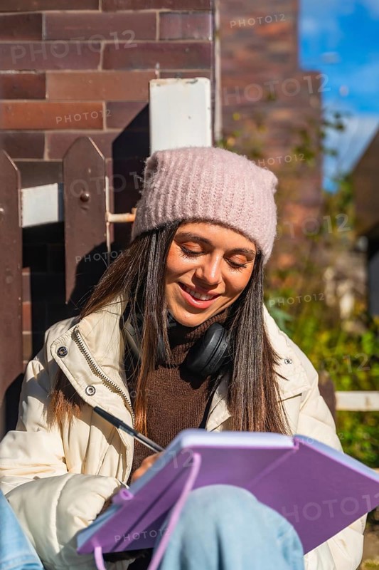 Young woman in a beanie and headphones writing in a notebook outdoors on a sunny day, smiling and focused as she journals, studies ideas, and enjoys a peaceful moment