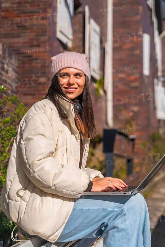 Woman in a cozy winter hat and jacket sitting outdoors, smiling while typing on a laptop, enjoying remote work and connectivity in an urban environment