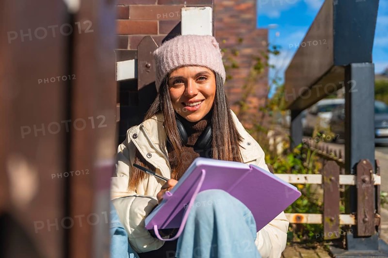 Young woman in beanie and headphones smiles while sketching in a notebook on a sunny urban street, relaxed and focused, enjoying creative outdoor downtime