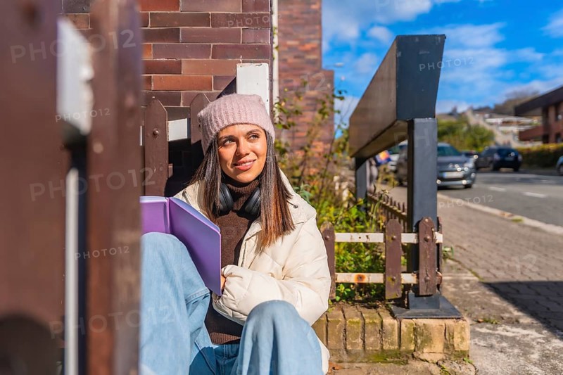 Young woman sitting outdoors on a sunny day, wearing a beanie and headphones, holding a book, looking away thoughtfully while enjoying a moment of relaxation and study