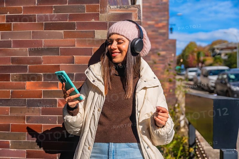 Woman smiling, wearing headphones and a beanie, holding a smartphone, standing against a brick wall on an autumn street, enjoying music and leisure time