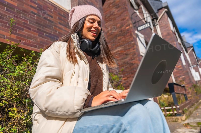 Young woman smiling and typing on a laptop, dressed in winter clothes, sitting outside a brick building on a sunny day, representing remote work and digital nomad lifestyle