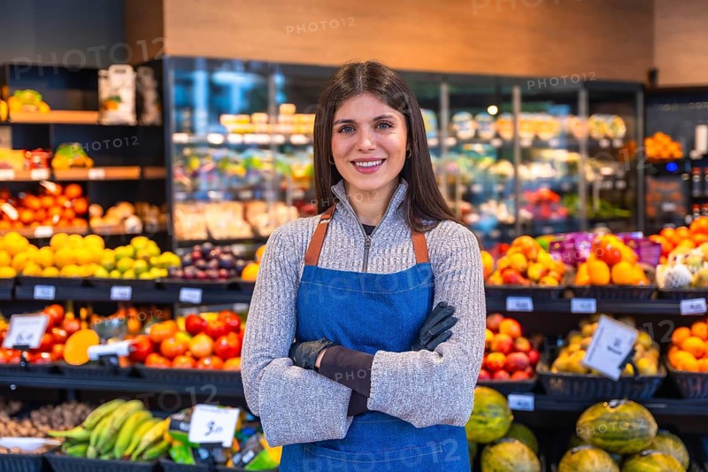 Young woman working in a fruit and vegetable section of a grocery store, wearing an apron and gloves, standing with arms crossed and smiling at the camera, promoting fresh produce