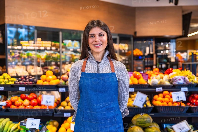 Woman wearing apron standing in a grocery store aisle, smiling at the camera, surrounded by fresh produce and fruits, representing retail business and customer service