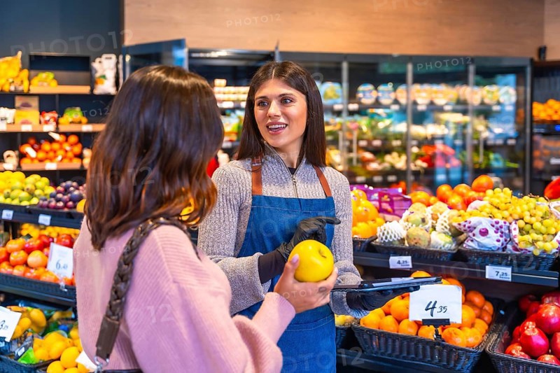 Supermarket employee wearing an apron and gloves helping a woman consumer select a fresh yellow apple from the fruit section, talking about product quality and service