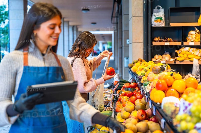 Greengrocer smiling and holding a digital tablet while a happy woman consumer chooses fresh tomatoes, fruits, and vegetables from an outdoor market stall, promoting healthy eating and local business