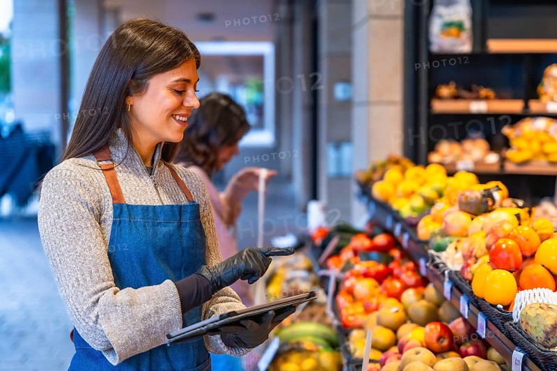 Female shop assistant wearing an apron and gloves managing fresh fruit and vegetables inventory with a digital tablet in a local grocery store, smiling while working