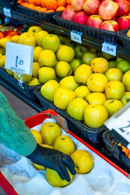 Person wearing a black glove carefully choosing ripe yellow apples from a display case at the fresh produce section in a supermarket, promoting healthy eating and conscious shopping choices