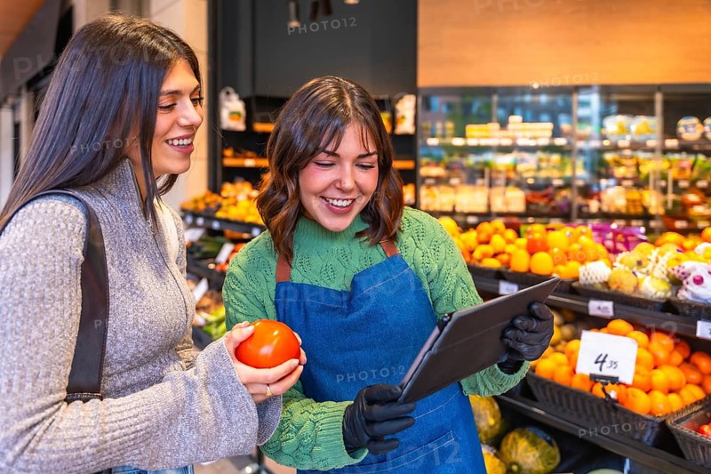 Store employee in an apron and gloves using a tablet while helping a smiling customer holding a fresh tomato in the vibrant produce section of a modern grocery store