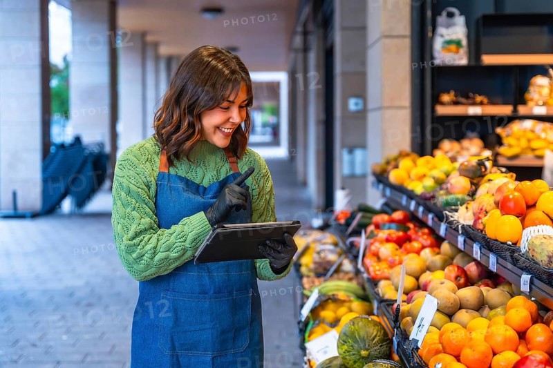 Woman in an apron and gloves managing fresh produce at her small business, using a digital tablet for inventory control while standing next to vibrant displays of fruit and vegetables