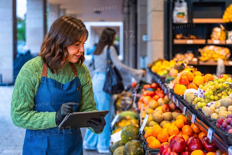 Woman in an apron and gloves managing inventory, checking stock levels of fresh fruit and produce on a digital tablet in a small business grocery store with a customer in the background