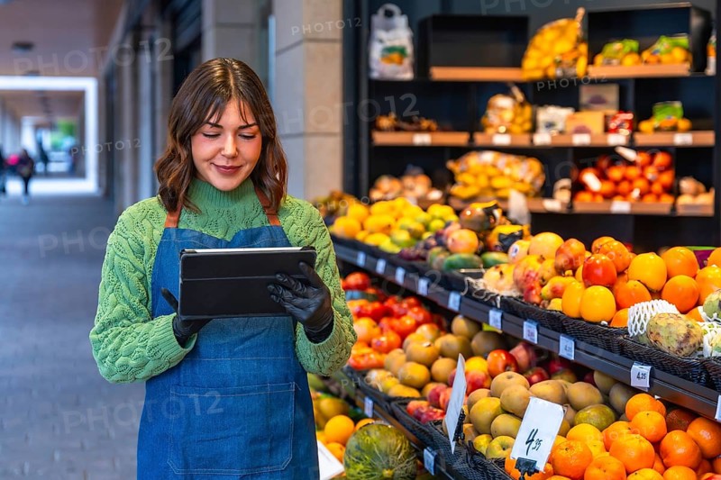 Female small business owner wearing an apron and gloves managing stock and orders on a digital tablet while standing in front of a colorful fruit and vegetable display