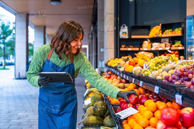 Smiling woman wearing an apron and gloves using a digital tablet for inventory management while restocking fresh fruits on display shelves in a small business produce shop