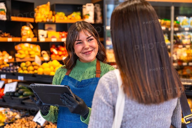 Friendly saleswoman holding a tablet and wearing gloves, assisting a customer with produce in a modern supermarket while offering excellent service and support