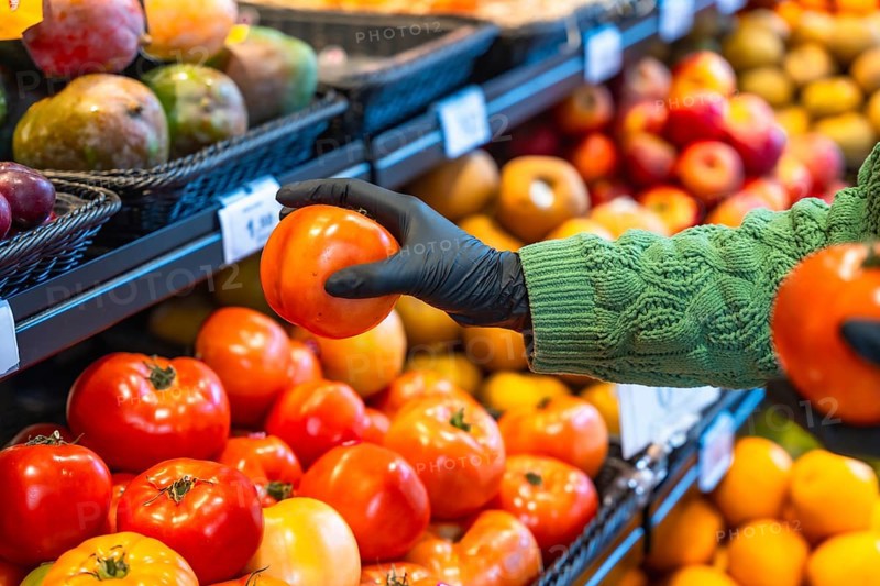 Person wearing a protective rubber glove selecting a ripe tomato from a produce display at a grocery store, representing safety during shopping and healthy eating