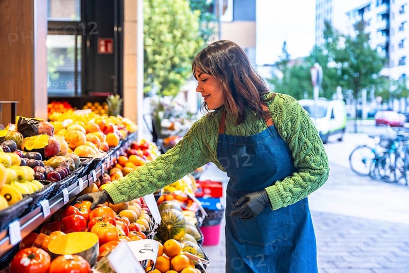 Smiling young greengrocer woman wearing an apron and gloves arranging fresh fruit and vegetables on market stalls outside her small business, ensuring quality and presentation