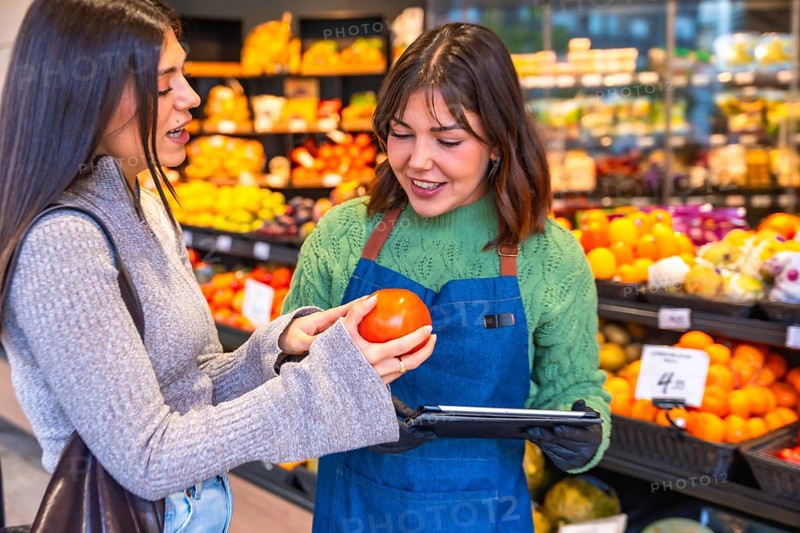 Grocery store employee in an apron holding a tablet and helping a female customer examine a fresh tomato in the produce aisle, fostering excellent customer service and shopping experience