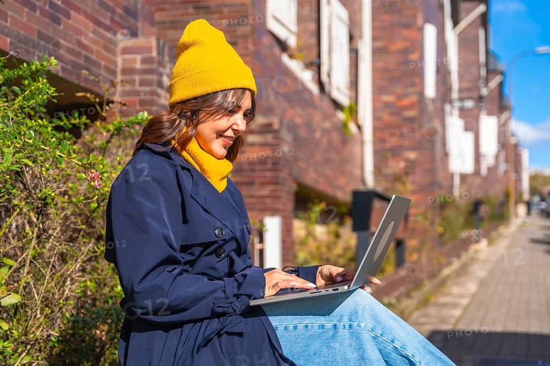 Smiling woman wearing a yellow beanie and blue trench coat, typing on a laptop while sitting outdoors in an urban environment, embracing flexible remote work and digital nomad lifestyle
