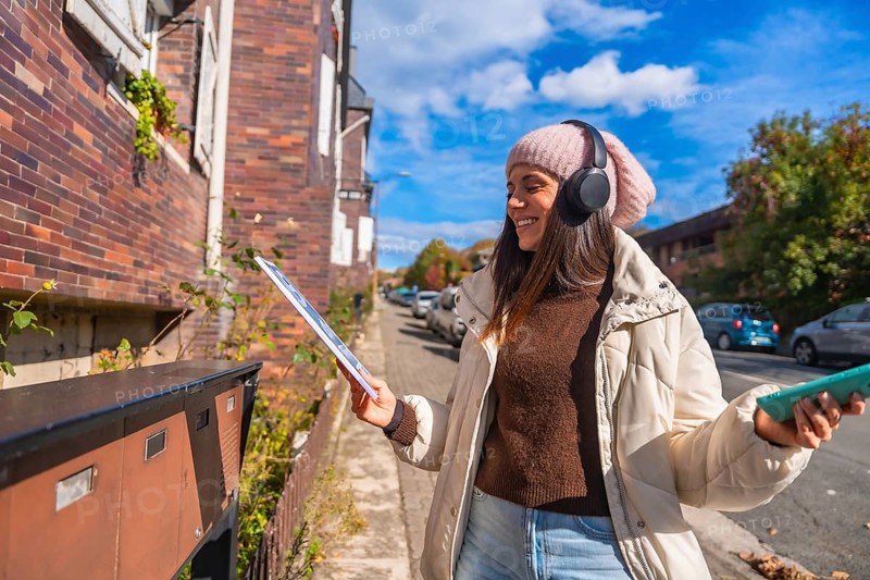Happy young woman standing on an urban street, opening a mailbox to retrieve mail while wearing headphones outdoors on a clear autumn day, enjoying her everyday routine
