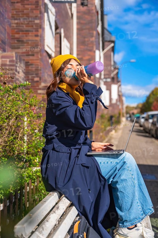 Woman hydrating with a water bottle while working remotely on her laptop, sitting on a bench in an urban setting during an autumn day, symbolizing flexible work and connection