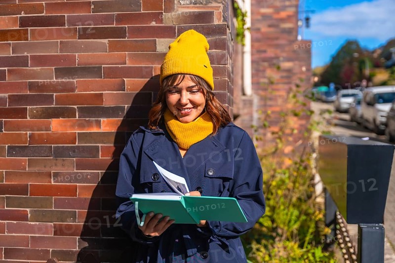 Young woman dressed in a trench coat and yellow beanie enjoying journaling and creative writing in her notebook during a sunny autumn day on an urban street