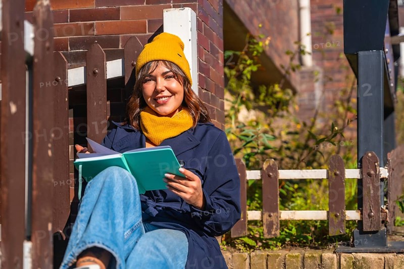 Young woman wearing a yellow beanie and scarf. Sitting outdoors in autumn sunlight. Smiling while reading a journal and looking at the camera. Capturing a moment of personal reflection and relaxation