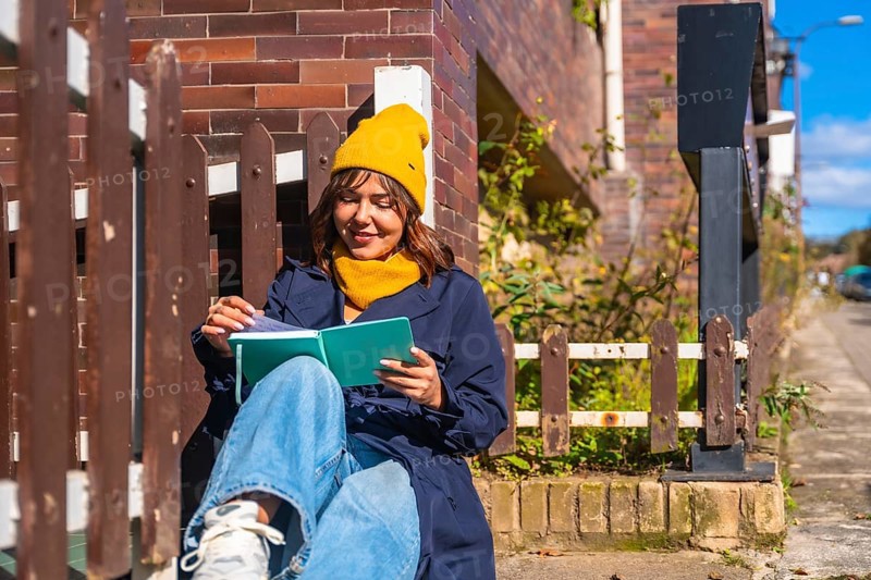 Woman wearing a yellow beanie and blue coat sitting on steps, enjoying a book in the warm sunlight by a brick building and fence, finding a moment of urban relaxation and quiet time