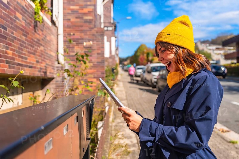 Young smiling woman wearing a yellow beanie and blue trench coat showing excitement finding a letter in an outdoor street mailbox, illustrating communication, surprise, and positive emotion
