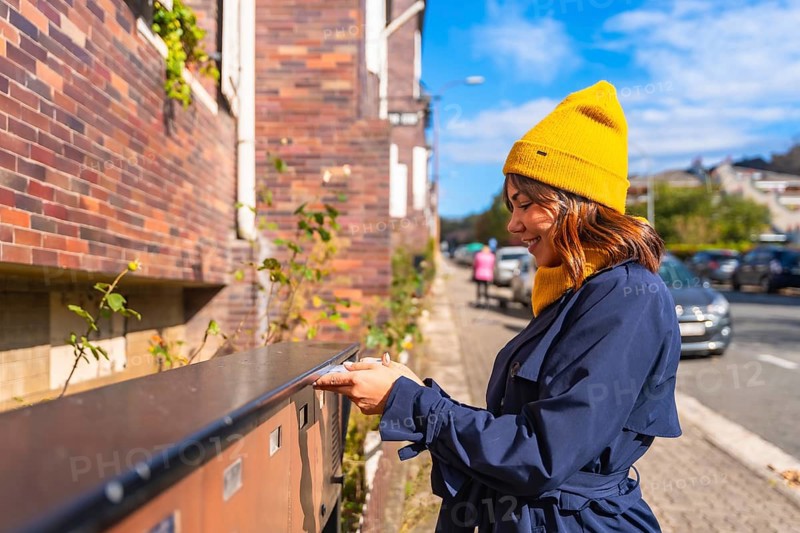 Woman wearing a yellow beanie and blue trench coat, smiling while sending a letter through a street mailbox, connecting with others through traditional mail in an urban setting