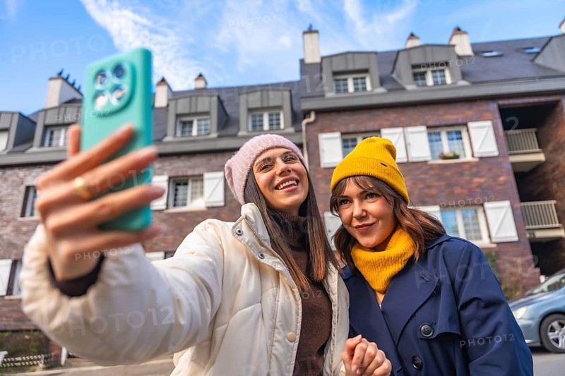 Two young women friends enjoying a fun moment together, smiling and taking a selfie outdoors in a city street during winter, wearing warm beanies and coats