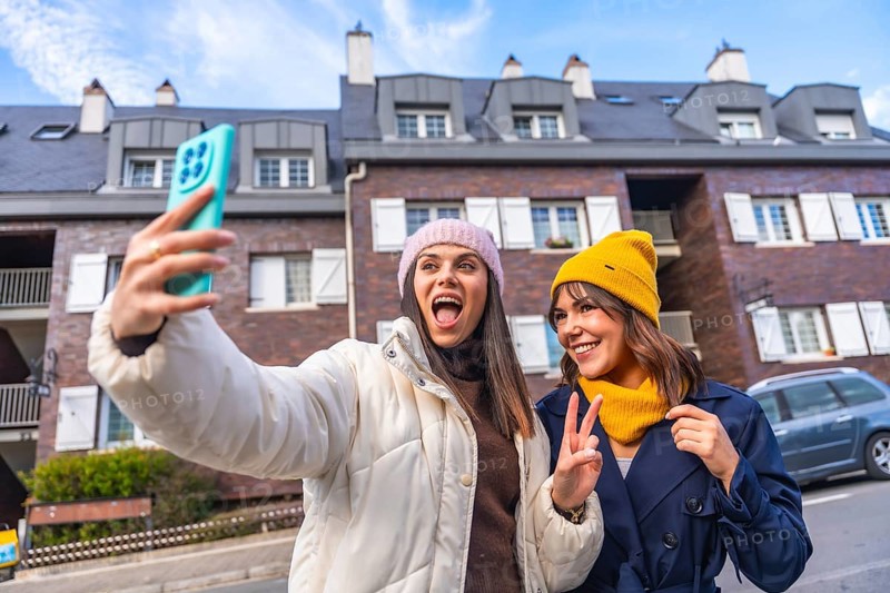 Two cheerful young women take a sunny winter selfie on a city street, bundled in beanies and jackets, smiling and capturing a fun travel moment together