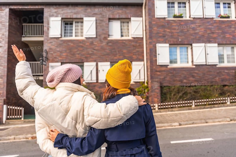 Two young women walking arm in arm down a city sidewalk in cool weather, wearing parkas and beanies, sharing a cheerful moment of friendship and connection during a casual stroll