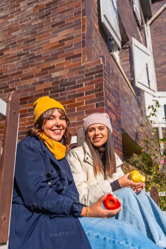 Two friends in warm winter clothes sit against a brick wall, smiling and sharing red and yellow apples on a sunny day, enjoying a casual, healthy moment together