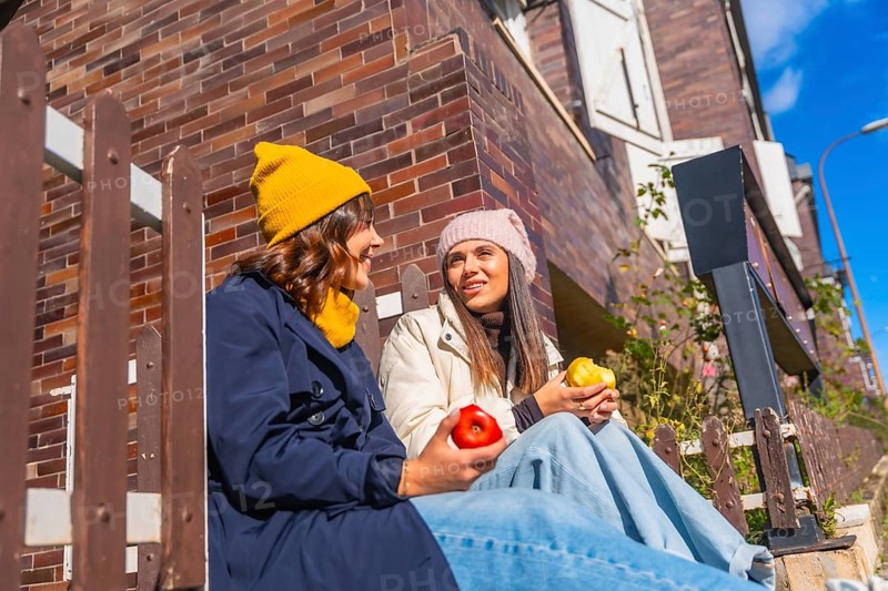Two young women sit on outdoor steps by a brick wall under blue sky, smiling and chatting while enjoying apples, cozy in beanies and coats on a sunny autumn day