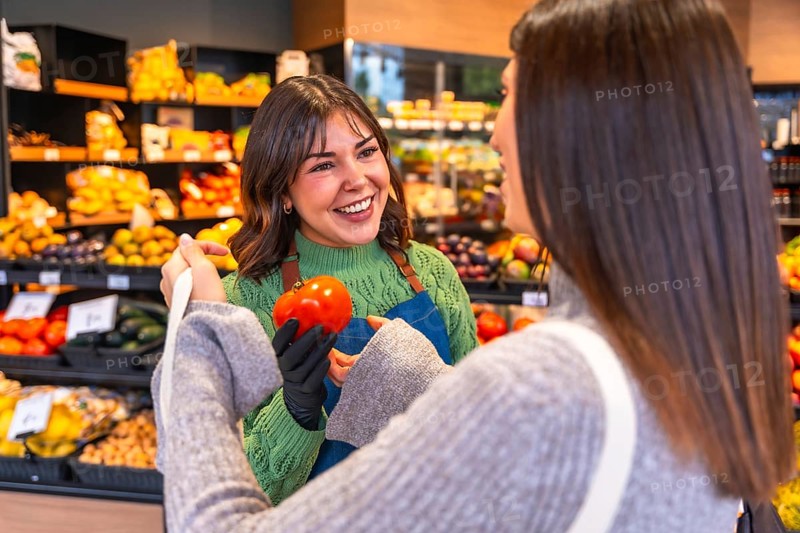 Friendly grocery store employee wearing an apron and gloves, showing a fresh red tomato to a smiling female customer, offering helpful service in the produce aisle