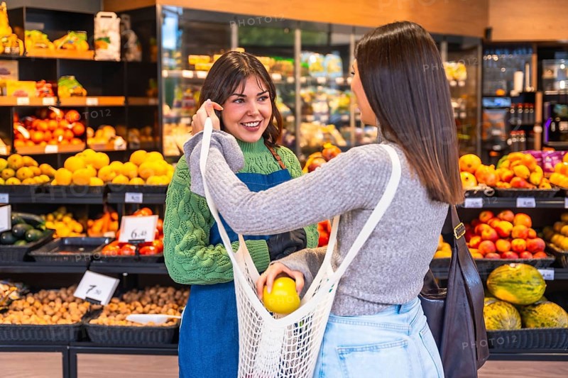 Friendly supermarket employee smiling and assisting a customer holding a reusable net bag to put fresh organic fruit in, promoting sustainable shopping and good service in a grocery store