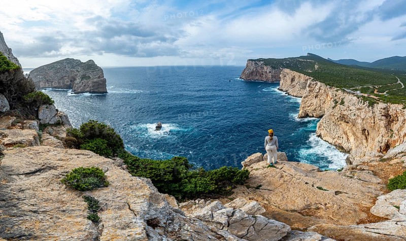 Tourist enjoying the view of steep cliffs by the sea and the rocky island of Isola Foradada, cliffs on the Capo Caccia headland, Alghero, Sardinia, Italy