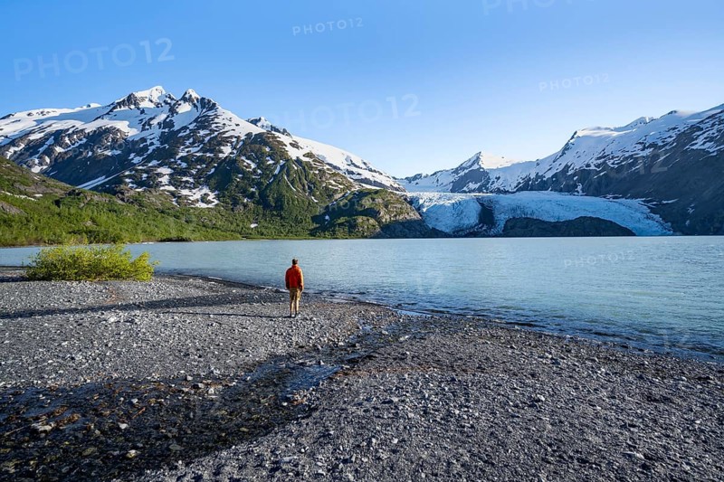 Young man on a pebble beach on a glacial lake, snowy mountains and Portage Glacier glacier on Portage Lake glacial lake, Chugach National Forest, Alaska, USA